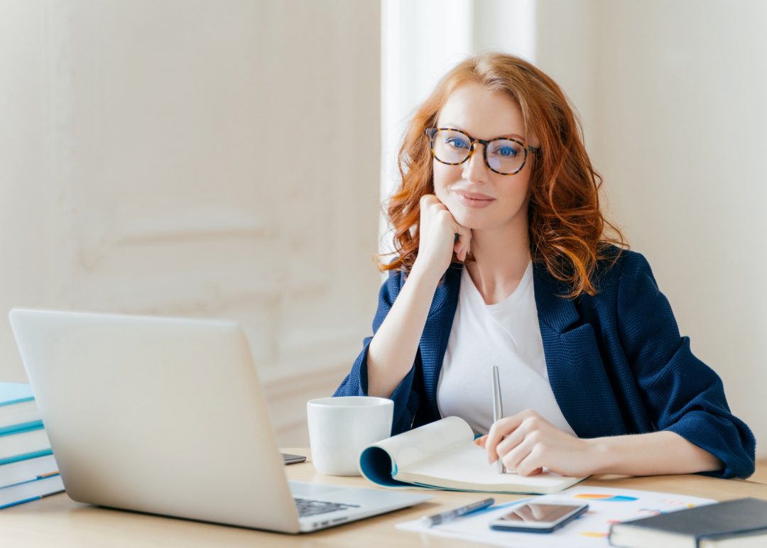 redhead-curly-woman-office-worker-analyzes-data-makes-accounting-report-poses-in-coworking-office